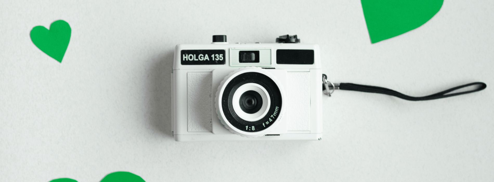 A white DSLR camera sits on a grey backdrop surrounded by green hearts