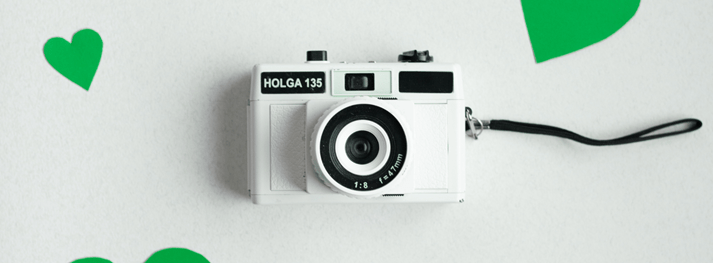 A white DSLR camera sits on a grey backdrop surrounded by green hearts